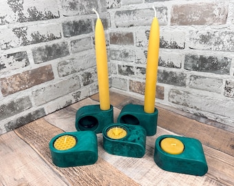 May include: Two tall yellow taper candles in dark green rounded holders and three low-profile dark green candle holders containing small yellow tea light candles with a honeycomb texture on the wax surface of two of the tea lights.
