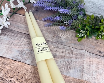 May include: Two pale yellow taper candles bound together by a cream-coloured paper label. The label features a bee icon and the text: Bees HANDMADE CANDLES 100% PURE BEESWAX. The candles have a smooth, cylindrical shape with a slightly tapered base.