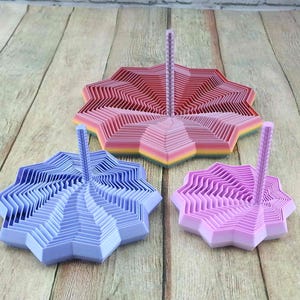 May include: Three decorative, star-shaped holders in shades of pink, purple, and blue. Each holder has a central, vertical post. The holders are arranged on a wooden surface, showcasing their layered, geometric design.