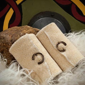 May include: Two beige fabric wrist cuffs with a herringbone pattern and bronze-coloured clasps. The cuffs are displayed on a bed of white feathers, with a large brown rock and a decorative shield in the background.