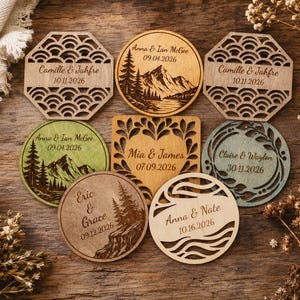 May include: A collection of seven circular and octagonal wooden ornaments featuring laser-engraved names and dates. Designs include mountain landscapes, geometric wave patterns, and leaf motifs. The pieces are crafted from light and stained wood.