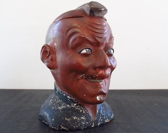 May include: A ceramic bust of a man with a painted face. The bust is painted in shades of red, brown, and black. The man has a painted mustache and a painted expression on his face. The bust is a vintage collectible.