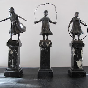 Op de afbeelding: Drie bronzen sculpturen van meisjes die met speelgoed spelen. Het meisje links houdt haar armen uitgestrekt, het meisje in het midden springt touwtje en het meisje rechts houdt een hoepel vast.