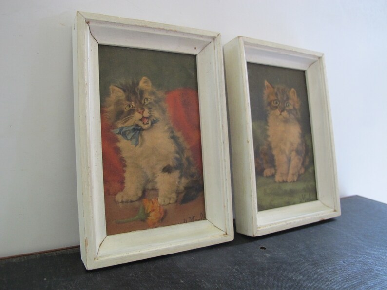 May include: Two framed pictures of kittens. The picture on the left shows a kitten with a blue ribbon around its neck. The picture on the right shows a kitten sitting on a green surface.