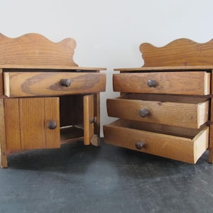 Puede incluir: Dos armarios de madera con cajones y puertas abiertos. El armario de la izquierda tiene un cajón y una puerta, mientras que el armario de la derecha tiene tres cajones. Ambos armarios tienen una parte superior decorativa.