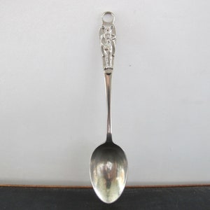 May include: Silver spoon with a detailed figure of a person on the handle. The spoon has a rounded bowl and a simple design.
