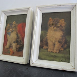 May include: Two framed pictures of kittens. The kitten in the left picture is holding a blue ribbon in its mouth. The kitten in the right picture is looking up. Both pictures have a white frame and a white mat.
