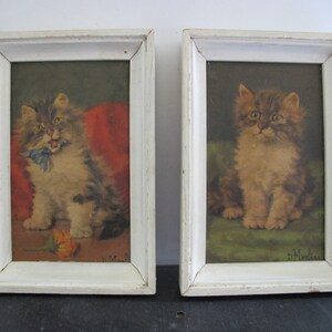 May include: Two framed paintings of kittens. The kitten on the left is sitting on a red cushion and has a blue ribbon around its neck. The kitten on the right is sitting on a green cushion. Both paintings are signed "H. Morris".