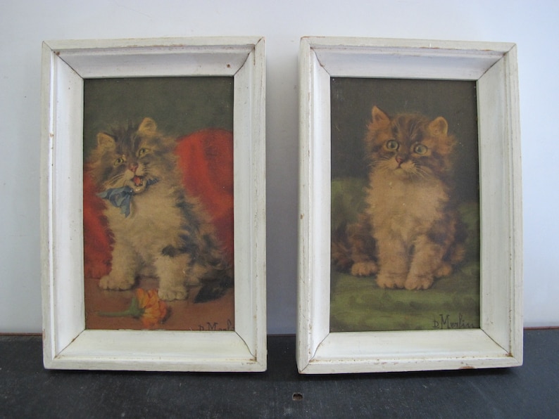 May include: Two framed paintings of kittens. The kitten on the left is sitting on a red cushion and has a blue ribbon around its neck. The kitten on the right is sitting on a green cushion. Both paintings are signed "H. Morris".