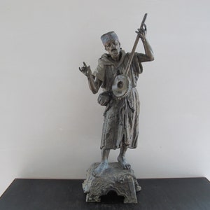 May include: A bronze sculpture of a man playing a musical instrument. The man is wearing a robe and a hat. The sculpture is on a wooden base.