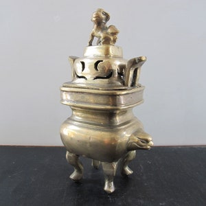 May include: A brass incense burner with a small lion figurine on top. The burner has a square base, curved legs, and decorative cutouts. The overall design is ornate and detailed, with a vintage aesthetic.