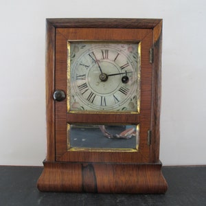 Puede incluir: Reloj antiguo de madera con una esfera blanca y marcadores de hora romanos negros. El reloj tiene una puerta de vidrio y un pequeño espejo debajo de la esfera.