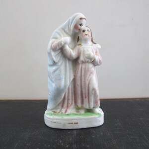 Puede incluir: Una figurita de porcelana de una mujer con una túnica azul y blanca que sostiene a una mujer más pequeña con un vestido rosa. La figurita está sobre una base verde.