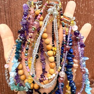 Puede incluir: Una mano muestra una colecci&oacute;n de coloridos collares de piedras preciosas. Los collares presentan varias cuentas, incluyendo amatista, lapisl&aacute;zuli y cuentas de madera. La joyer&iacute;a es en tonos p&uacute;rpura, azul, rosa y dorado.