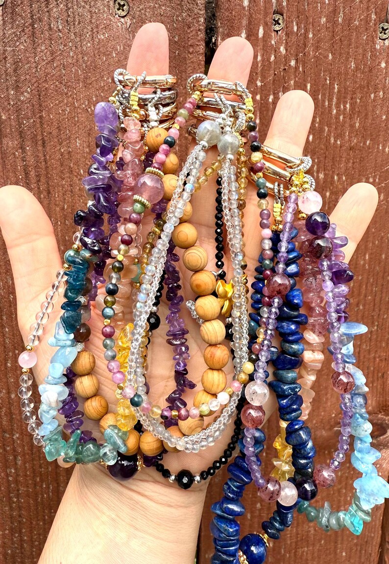 Puede incluir: Una mano muestra una colecci&oacute;n de coloridos collares de piedras preciosas. Los collares presentan varias cuentas, incluyendo amatista, lapisl&aacute;zuli y cuentas de madera. La joyer&iacute;a es en tonos p&uacute;rpura, azul, rosa y dorado.