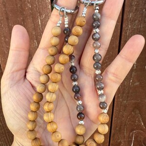 Puede incluir: Tres llaveros con cuentas de madera y piedra. Los llaveros se sostienen en una mano, mostrando los diferentes tipos de cuentas y anillos de metal plateado. Las cuentas de madera son de color marr&oacute;n claro, mientras que las cuentas de piedra son de tonos grises y marrones.