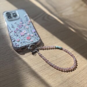 Puede incluir: Funda de tel&eacute;fono transparente con un dise&ntilde;o floral en rosa y morado. La funda est&aacute; sujeta a una correa de mu&ntilde;eca con cuentas rosas y un peque&ntilde;o acento turquesa. La funda del tel&eacute;fono est&aacute; sobre una superficie de madera.