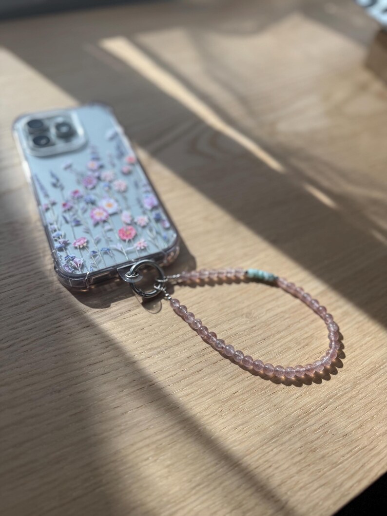 Puede incluir: Funda de tel&eacute;fono transparente con un dise&ntilde;o floral en rosa y morado. La funda est&aacute; sujeta a una correa de mu&ntilde;eca con cuentas rosas y un peque&ntilde;o acento turquesa. La funda del tel&eacute;fono est&aacute; sobre una superficie de madera.