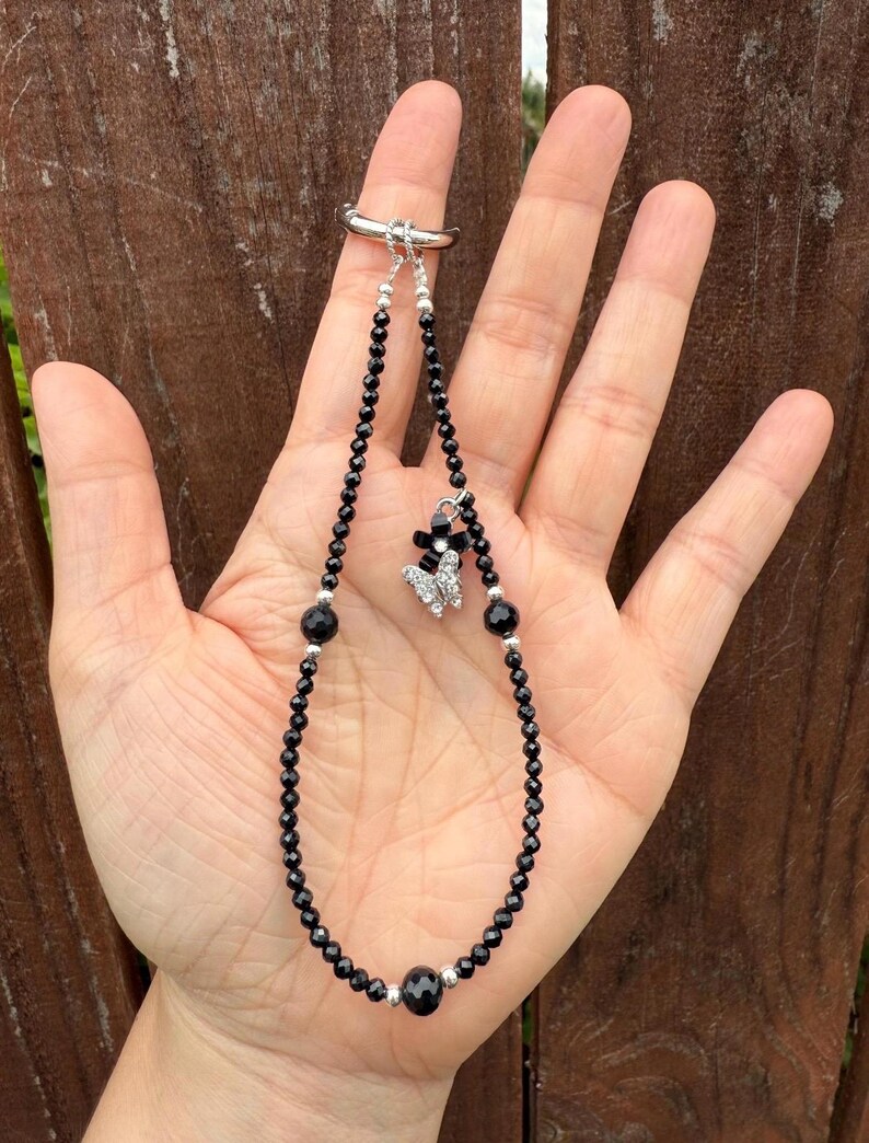 Puede incluir: Una pulsera de cuentas negras con un enganche de anillo plateado. La pulsera presenta un dije de mariposa con detalles en negro y plata. Las cuentas son peque&ntilde;as y redondas, con cuentas negras m&aacute;s grandes intercaladas. La pulsera se sujeta con un anillo plateado.