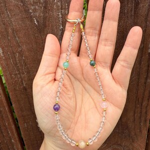 Puede incluir: Un delicado collar de cuarzo transparente con detalles dorados y cuentas de piedras preciosas de colores. El collar presenta un cierre de anillo dorado y se muestra sobre un fondo de madera.