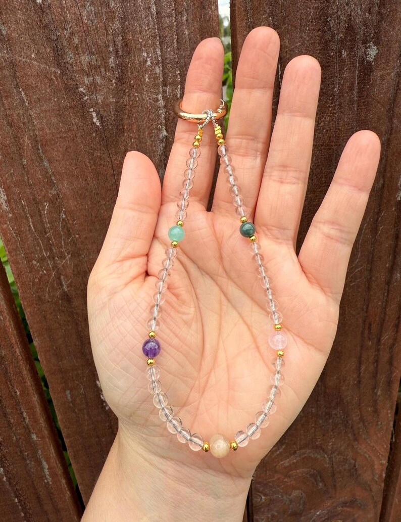 Puede incluir: Un delicado collar de cuarzo transparente con detalles dorados y cuentas de piedras preciosas de colores. El collar presenta un cierre de anillo dorado y se muestra sobre un fondo de madera.