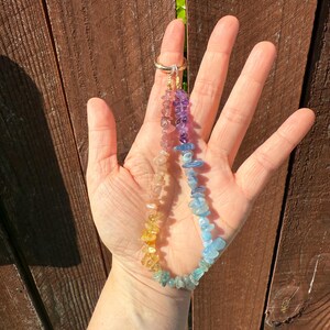 Puede incluir: Un collar de piedras preciosas de colores del arco&iacute;ris con un cierre de anillo dorado. El collar presenta un degradado de colores, incluyendo amarillo, naranja, rosa, morado y azul. Las piedras preciosas son peque&ntilde;as, de formas irregulares. El collar se sostiene en una mano.