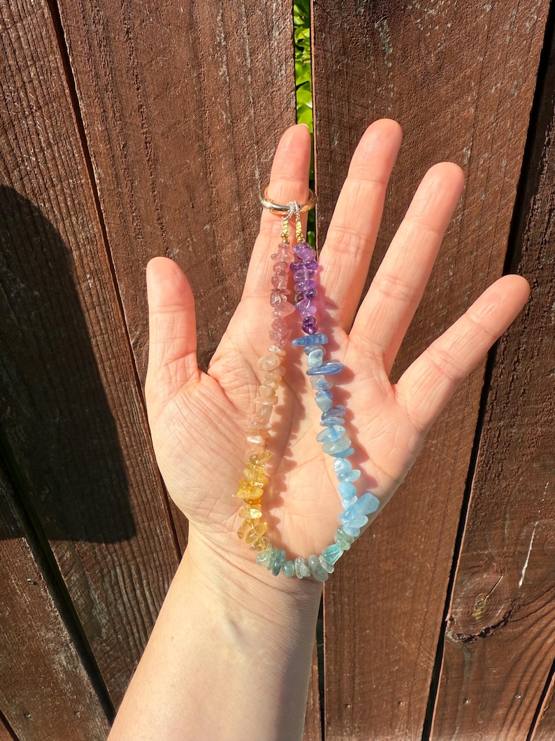 Puede incluir: Un collar de piedras preciosas de colores del arco&iacute;ris con un cierre de anillo dorado. El collar presenta un degradado de colores, incluyendo amarillo, naranja, rosa, morado y azul. Las piedras preciosas son peque&ntilde;as, de formas irregulares. El collar se sostiene en una mano.