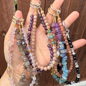 Puede incluir: Una mano muestra varias pulseras de piedras preciosas en tonos rosa, morado, azul y negro. Cada pulsera presenta una combinaci&oacute;n &uacute;nica de cuentas y cristales, con detalles dorados y plateados.