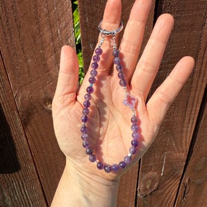 Puede incluir: Un llavero de cuentas con piedras preciosas de amatista. El llavero tiene un anillo plateado y un dije decorativo con una piedra rosa en forma de coraz&oacute;n. Las cuentas de amatista son de color p&uacute;rpura oscuro.