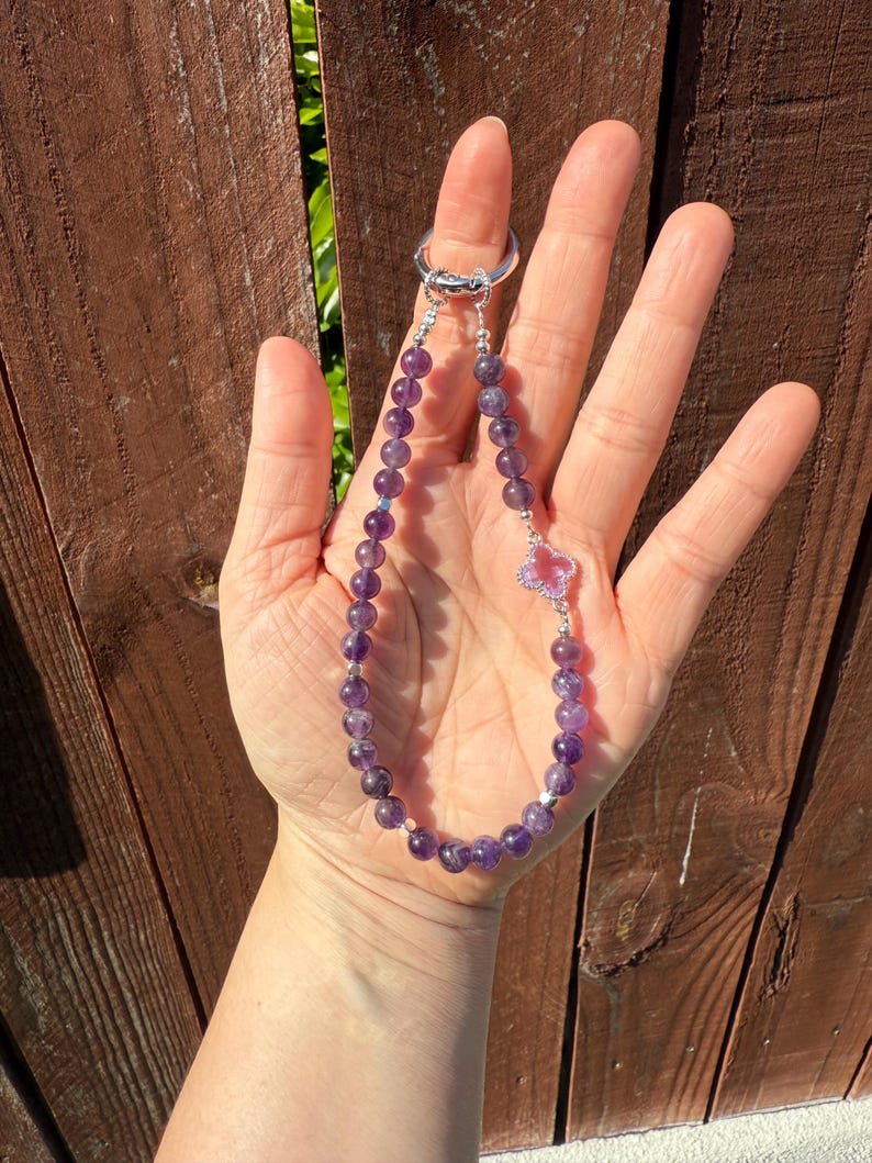 Puede incluir: Un llavero de cuentas con piedras preciosas de amatista. El llavero tiene un anillo plateado y un dije decorativo con una piedra rosa en forma de coraz&oacute;n. Las cuentas de amatista son de color p&uacute;rpura oscuro.