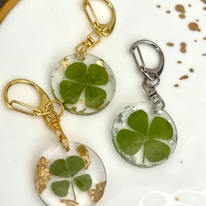 May include: Three clear resin keychains with four-leaf clovers encased inside. Two keychains have gold-colored hardware and gold leaf accents, while the third has silver-colored hardware and silver leaf accents. The keychains are round.