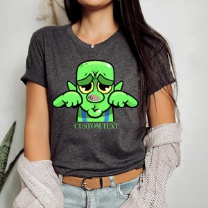 May include: Dark gray t-shirt featuring a cartoon green creature with a sad expression. The creature has large yellow eyes, a pink nose, and blue and green accents. The text "CUSTOM TEXT" is below the creature.
