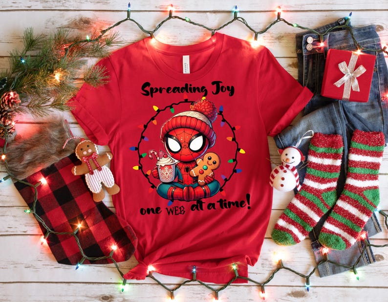 May include: Red t-shirt featuring a cartoon Spider-Man character in a winter hat, surrounded by Christmas lights. The shirt has the text "Spreading Joy" and "one WEB at a time!". The image includes a gingerbread man, a cup of cocoa, and Christmas decorations.