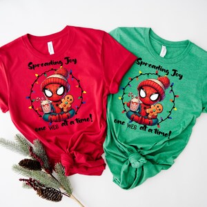 May include: Red and green t-shirts featuring a cartoon Spider-Man character in a winter hat, holding a gingerbread man and a drink. The shirts have the text "Spreading Joy one WEB at a time!" printed on them. Festive holiday apparel.
