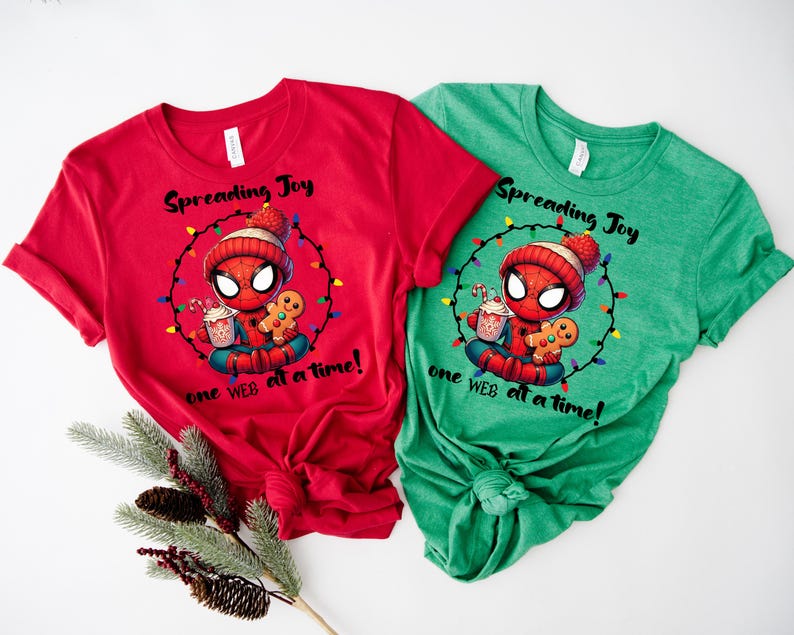 May include: Red and green t-shirts featuring a cartoon Spider-Man character in a winter hat, holding a gingerbread man and a drink. The shirts have the text "Spreading Joy one WEB at a time!" printed on them. Festive holiday apparel.