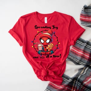 May include: Red t-shirt featuring a cartoon Spider-Man character in a winter hat, surrounded by Christmas lights. The graphic includes the text "Spreading Joy one web at a time!" and images of a gingerbread man and a cup of cocoa.
