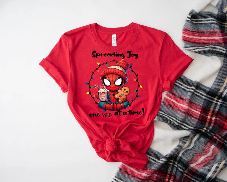 May include: Red t-shirt featuring a cartoon Spider-Man character in a winter hat, surrounded by Christmas lights. The graphic includes the text "Spreading Joy one web at a time!" and images of a gingerbread man and a cup of cocoa.