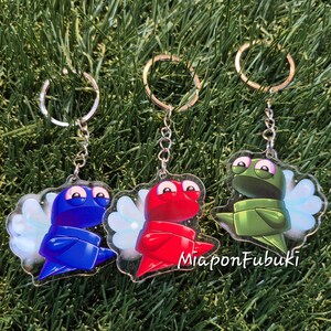 Peut inclure: Trois porte-clés en acrylique avec des personnages de dessins animés bleus, rouges et verts. Chaque personnage a de grands yeux, des ailes et une écharpe. Les porte-clés sont sur des chaînes argentées avec des anneaux. Le texte "MiaponFubuki" est visible.