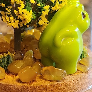 May include: A small terrarium featuring a bright green figurine, a miniature tree with yellow flowers, and amber-colored stones. The terrarium is enclosed in a clear glass dome, creating a miniature landscape.