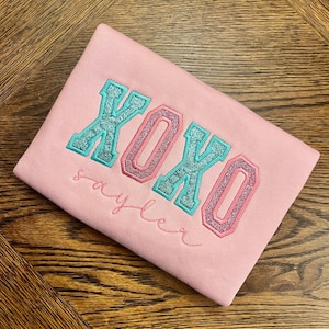 May include: Pink sweatshirt with the word "XOXO" in large, glittery, multi-colored letters. The word "Sayler" is written in cursive below the letters.