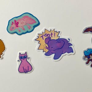 May include: A set of five colorful stickers featuring various animals. The stickers include a pink axolotl, a brown capybara, a purple elephant with the word "Toot!" in a yellow speech bubble, a pink cat, and a brown ferret wearing a blue hat.