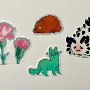 May include: A set of five stickers featuring a green cat, a brown bear, a pink flower, a white flower, and a black and white sea slug.