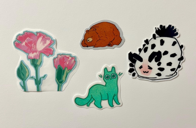 May include: A set of five stickers featuring a green cat, a brown bear, a pink flower, a white flower, and a black and white sea slug.