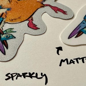 Peut inclure: Gros plan sur deux autocollants en forme d'oiseau. L'un est scintillant avec des couleurs orange, bleu et violet. L'autre est mat avec des couleurs similaires. Les mots "sparkly" et "matte" sont &eacute;crits &agrave; l'encre noire.