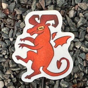 May include: A red cartoon dragon sticker with a white border. The dragon has horns, wings, and a curled tail. The sticker is placed on a bed of small gray rocks.