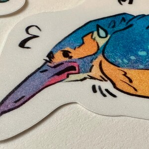 Peut inclure: Un autocollant repr&eacute;sentant une illustration stylis&eacute;e d'un martin-p&ecirc;cheur. L'oiseau est de couleur bleue, orange et violette avec des d&eacute;tails noirs. Le chiffre "3" est imprim&eacute; en noir sur l'autocollant.