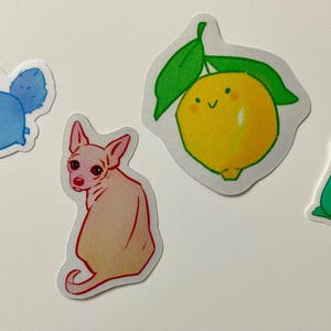 May include: Four colorful stickers featuring a blue cat with wings, a pink dog, a green pear, and a yellow lemon with a green stem and leaves.