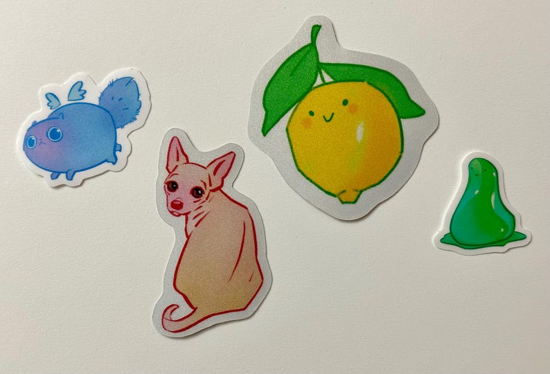 May include: Four colorful stickers featuring a blue cat with wings, a pink dog, a green pear, and a yellow lemon with a green stem and leaves.
