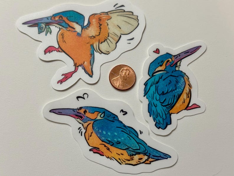 Peut inclure: Trois autocollants color&eacute;s de martin-p&ecirc;cheur avec une pi&egrave;ce de monnaie pour l'&eacute;chelle. Les autocollants pr&eacute;sentent des illustrations de l'oiseau dans diverses poses, avec des couleurs bleues, oranges et blanches. Un autocollant montre l'oiseau en vol, un autre avec un c&oelig;ur et le troisi&egrave;me avec des notes de musique.