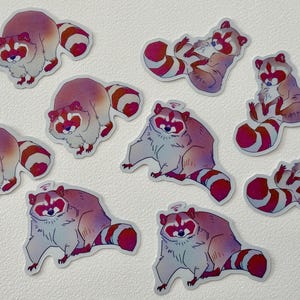 May include: Nine colorful raccoon stickers with pink, white, and gray fur. The raccoons are in various poses, including sitting, lying down, and standing. The stickers are perfect for adding a touch of whimsy to laptops, water bottles, or journals.